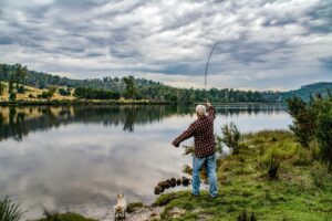Elderly man fishing by a picturesque lake in Ouse, Tasmania, with a dog by his side.